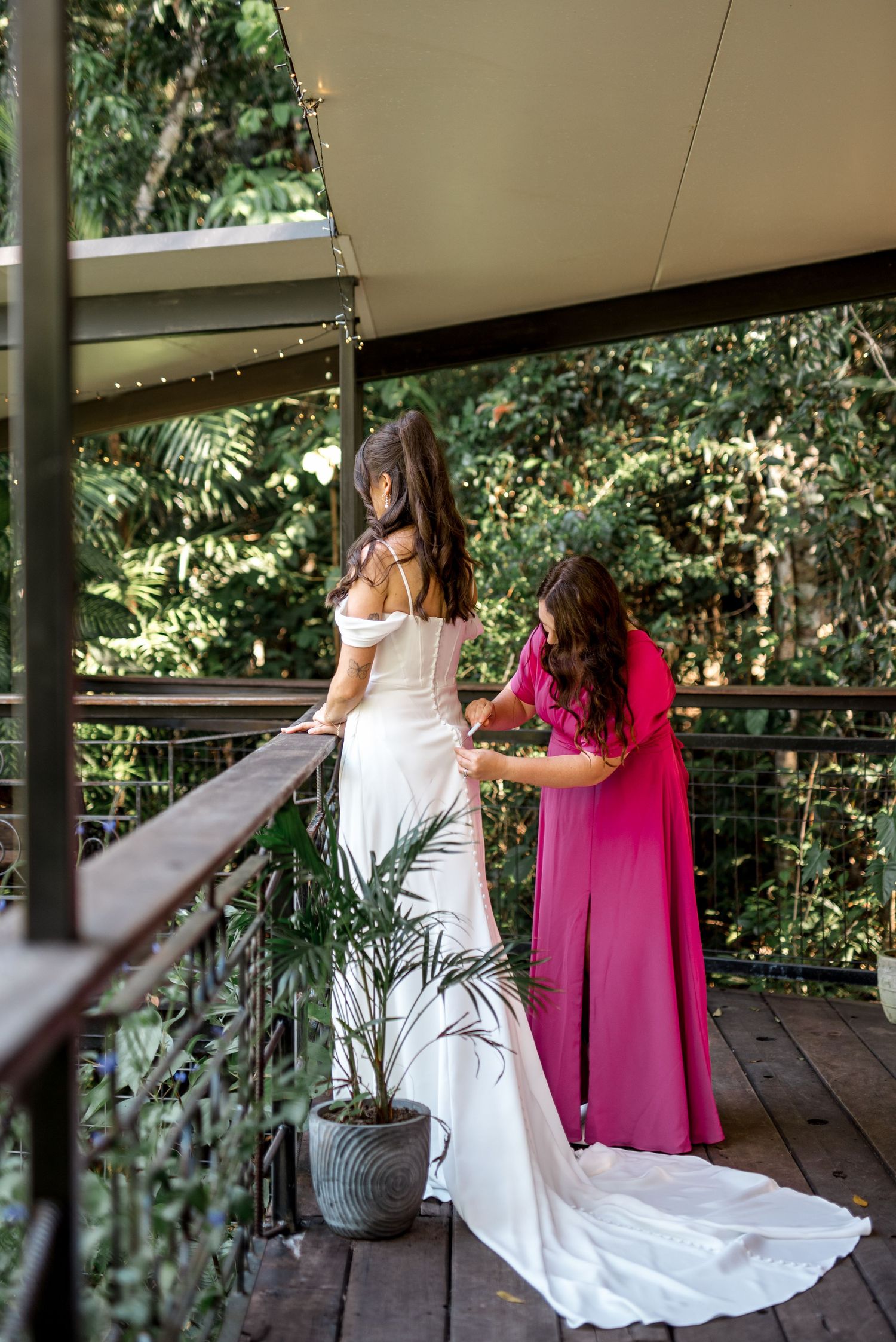 Bridesmaid in pink dress buttoning bride's white wedding gown on a wooden balcony surrounded by lush greenery.