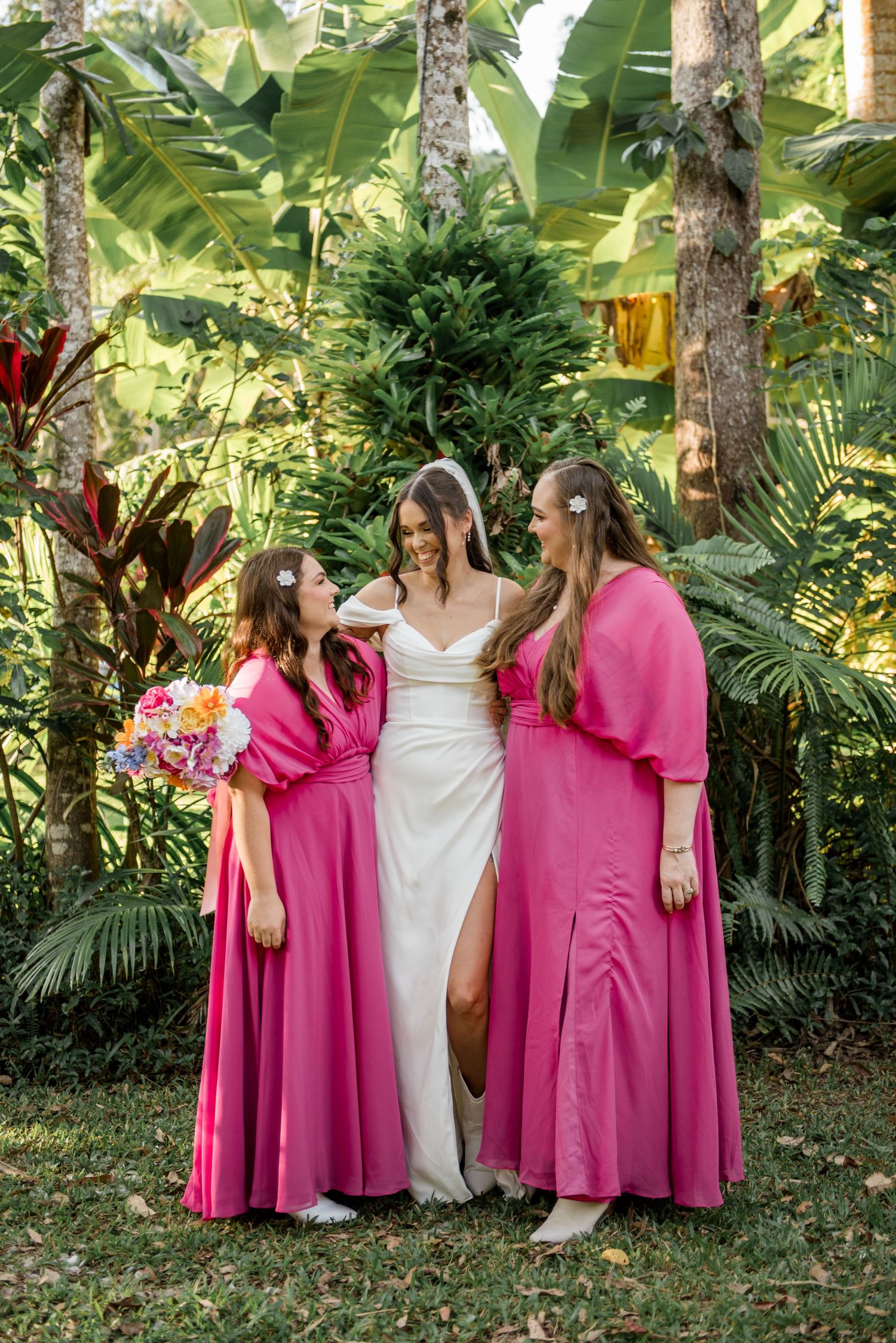 Bride in white gown smiles with two bridesmaids in hot pink dresses in tropical garden.