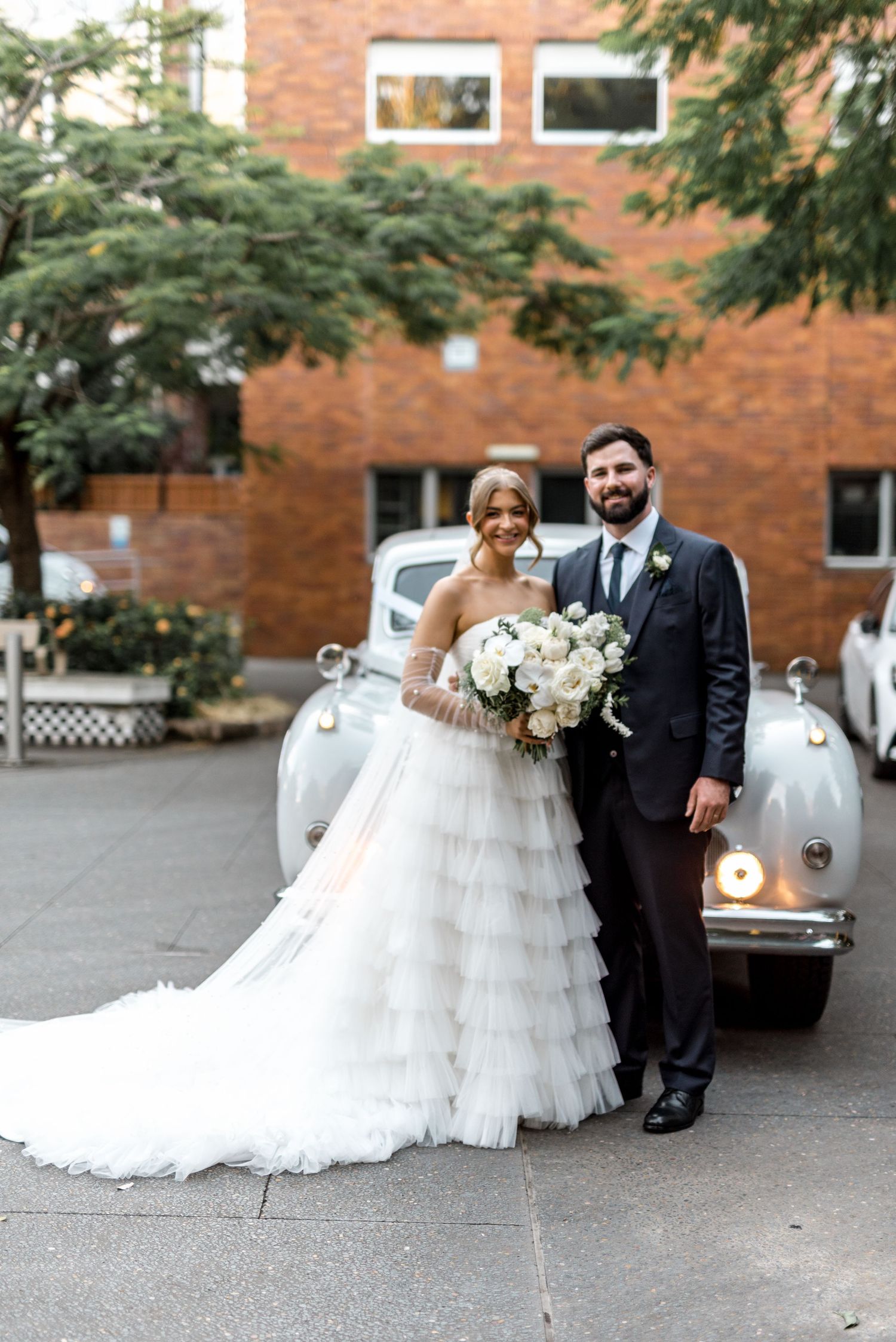 Smiling bride and groom pose in front of vintage white car, bride holding white floral bouquet.