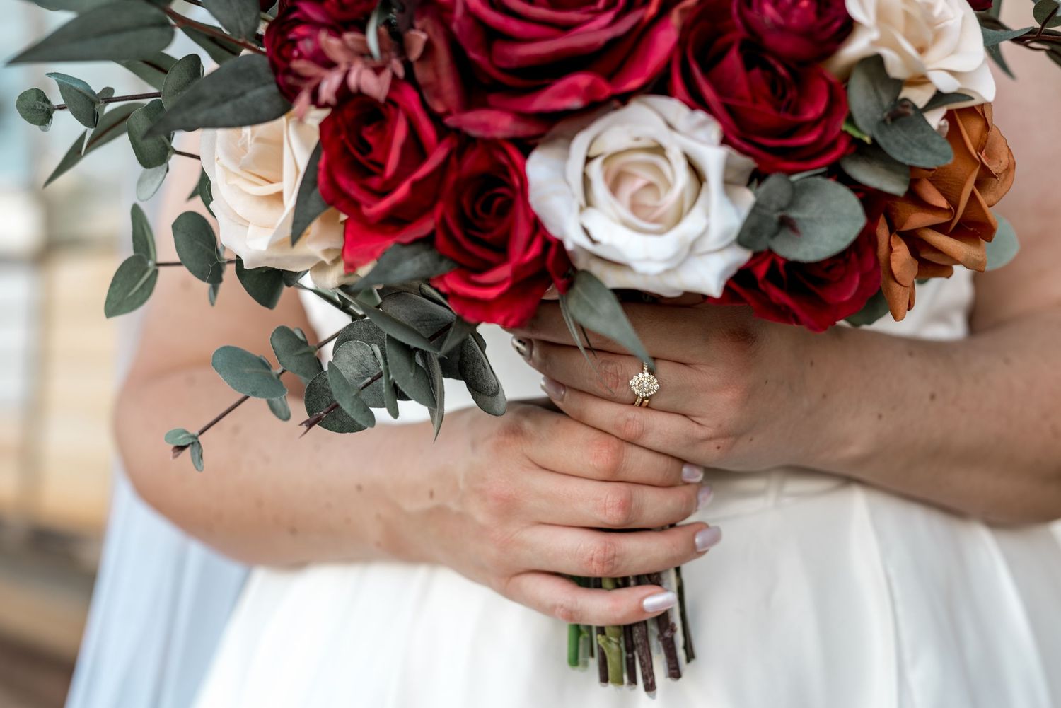 Bride holds a stunning bouquet of deep red and cream roses with eucalyptus and a diamond ring.