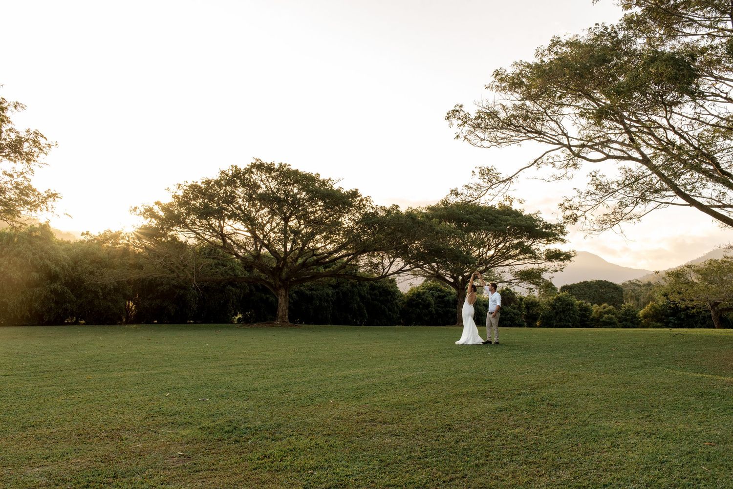 Bride and groom standing together on vast green lawn surrounded by trees at sunset.