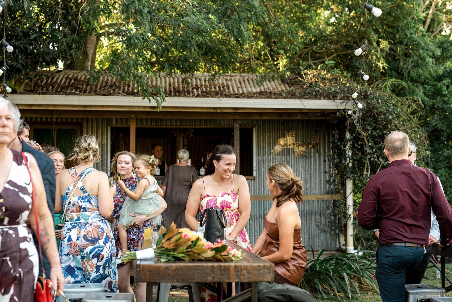 Wedding guests mingle around wooden table with tropical fruit at rustic outdoor reception.