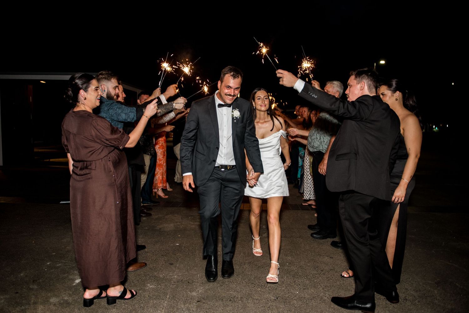 Smiling couple exits wedding reception through sparkler sendoff with guests lining path.