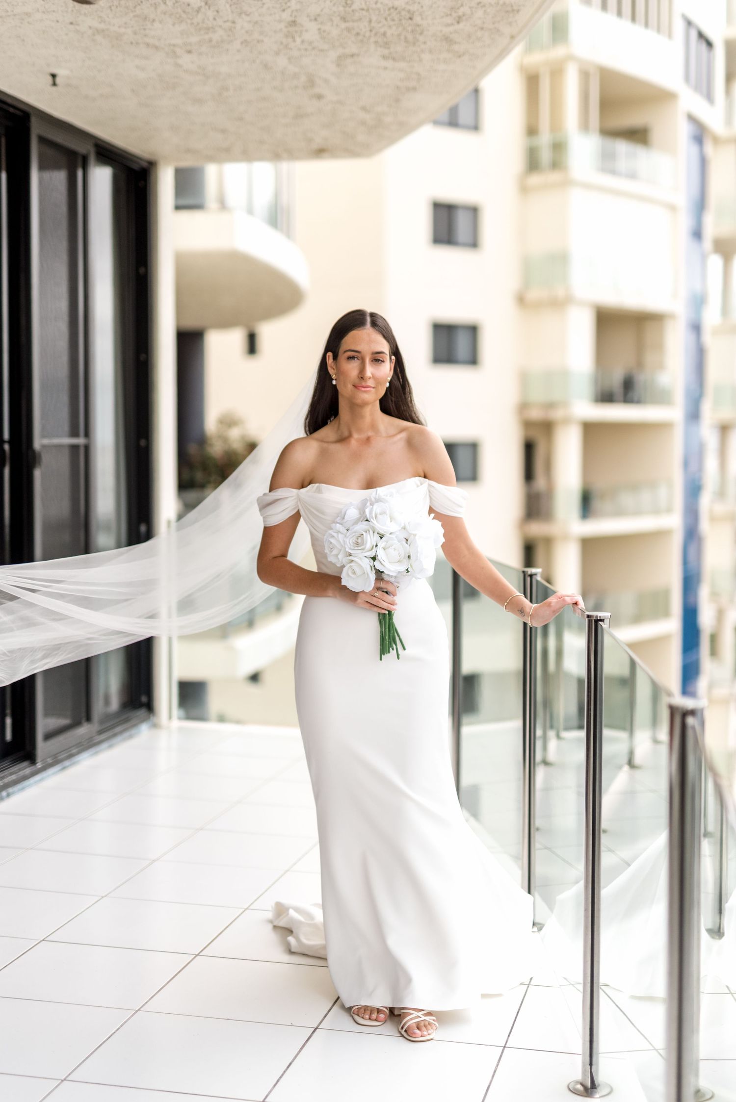Bride in white off-shoulder gown with flowing veil holding white roses on balcony.