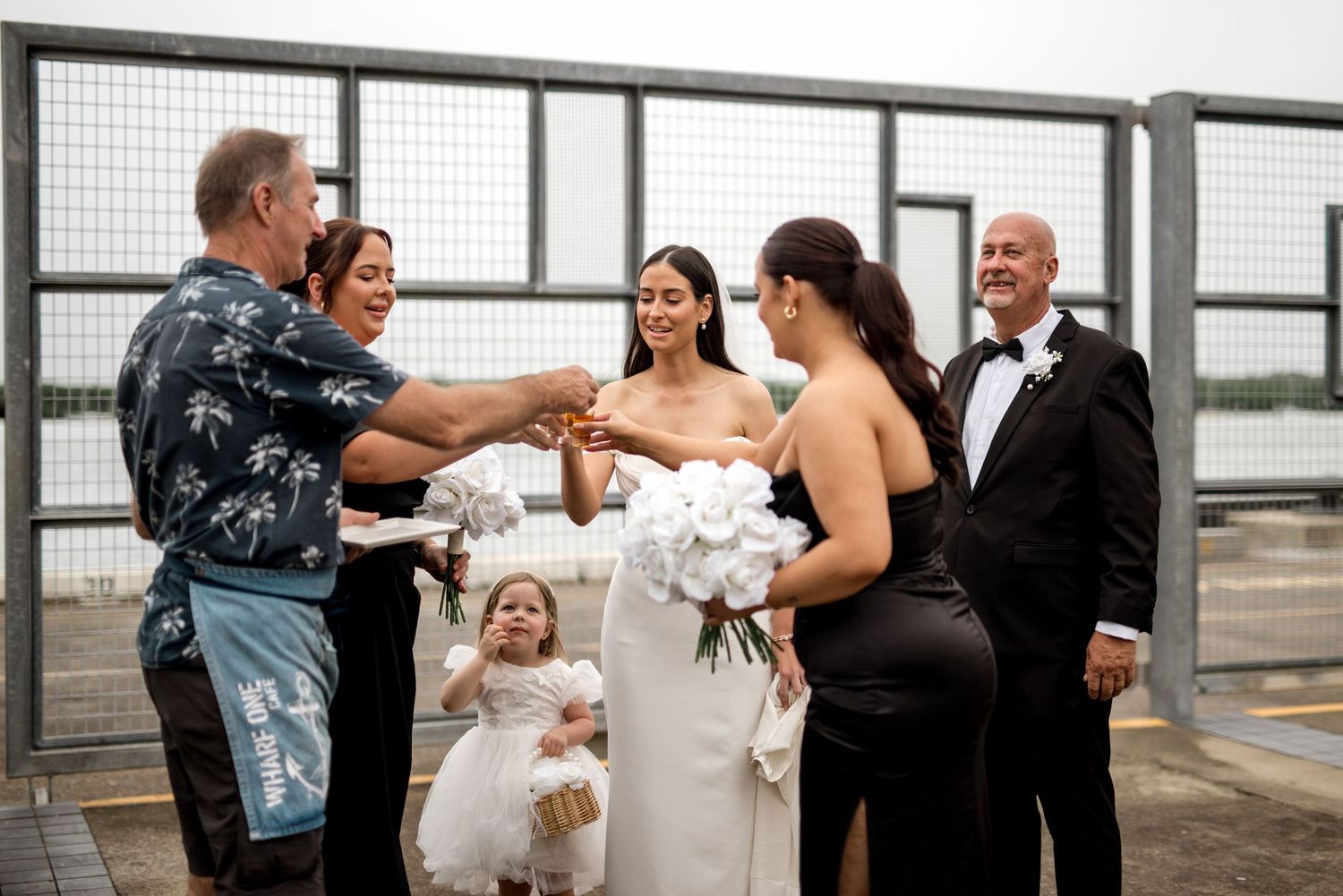 Wedding party laughing and celebrating outdoors near a metal fence by the water.