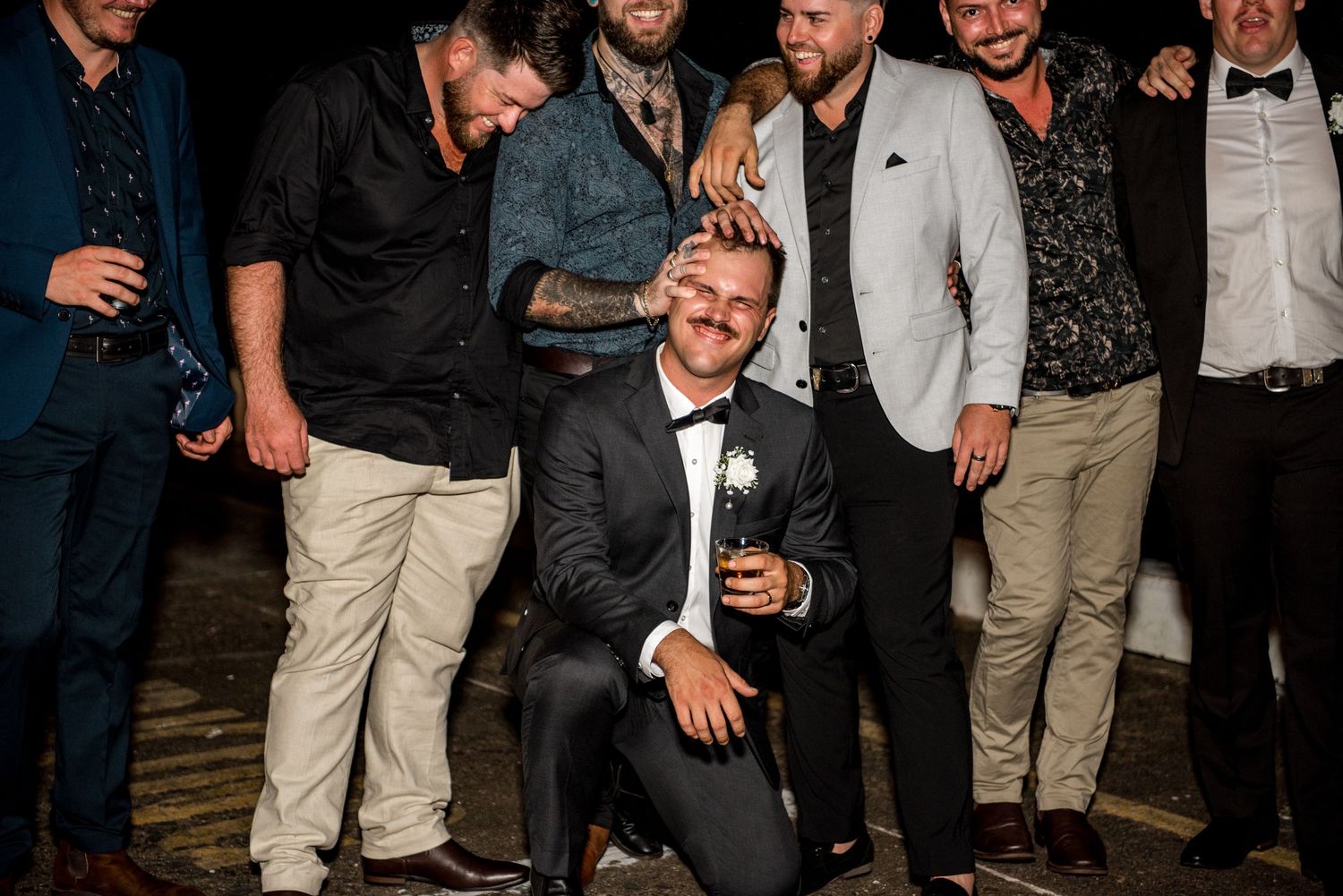 Groom kneels smiling surrounded by groomsmen laughing together at a wedding celebration.