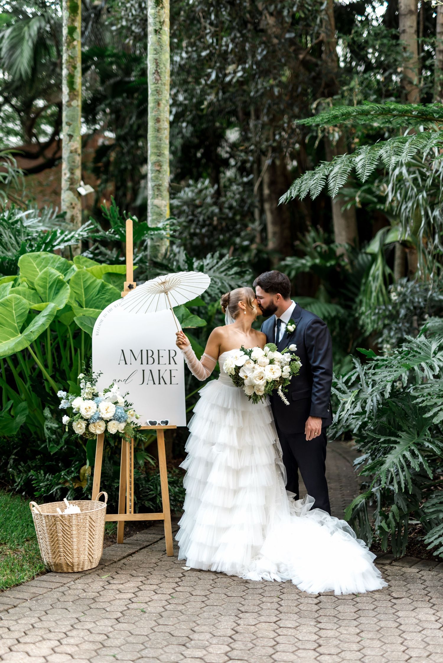 Bride and groom kiss beside Amber and Jake wedding welcome sign surrounded by tropical garden greenery.