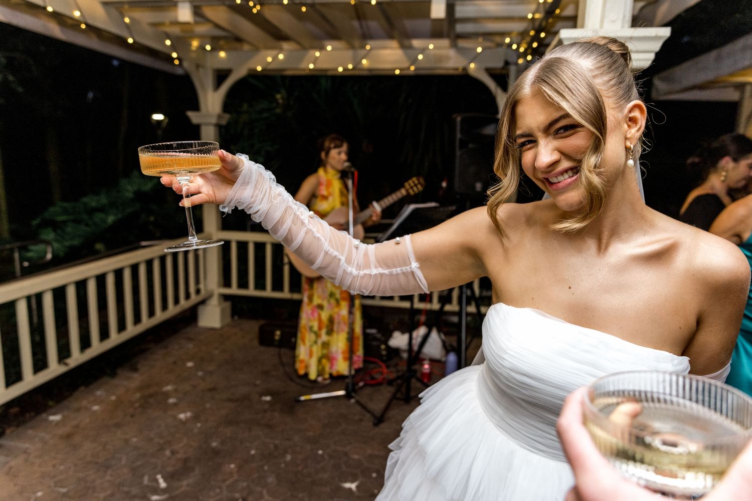 Smiling bride raises a champagne coupe glass at her outdoor wedding reception.