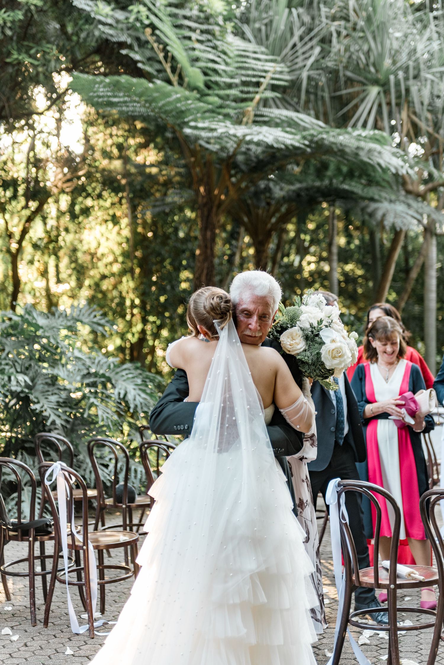 Bride in tiered white gown with long veil shares an emotional embrace with elderly guest during outdoor ceremony.