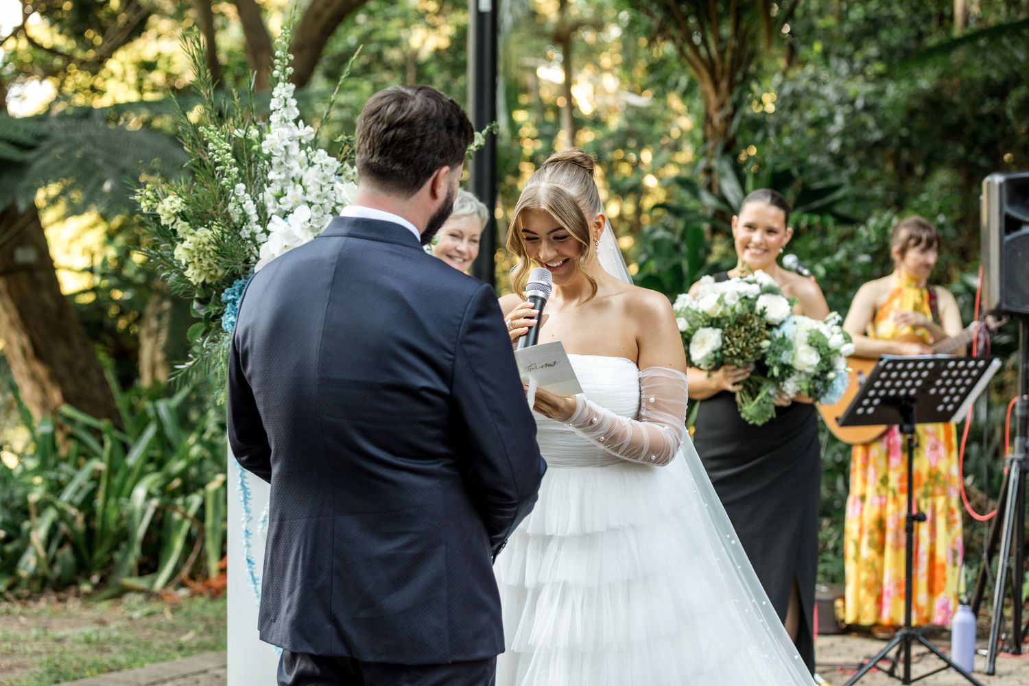 Smiling bride reciting vows into microphone during outdoor garden wedding ceremony.