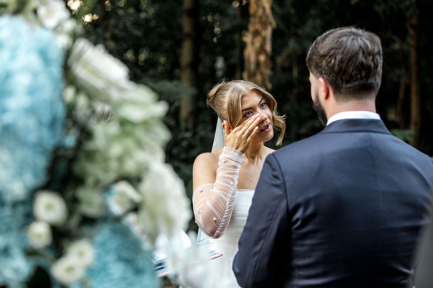 Emotional bride covers her mouth smiling at groom surrounded by blue and white floral arrangements.
