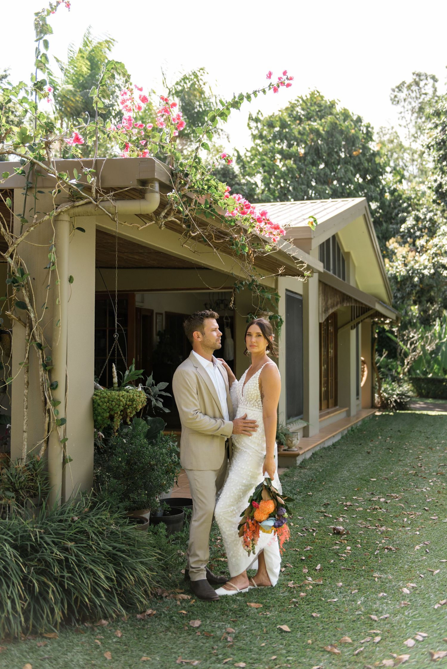 Bride holding tropical bouquet and groom in tan suit standing before a charming cottage with bougainvillea.