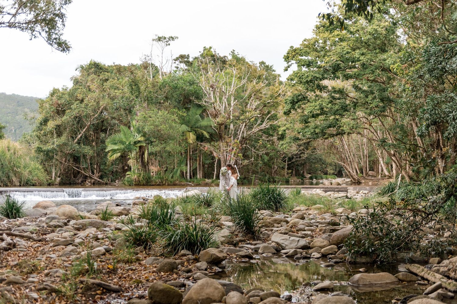 Wedding couple embrace in a lush green forest clearing surrounded by rocks and flowing water.