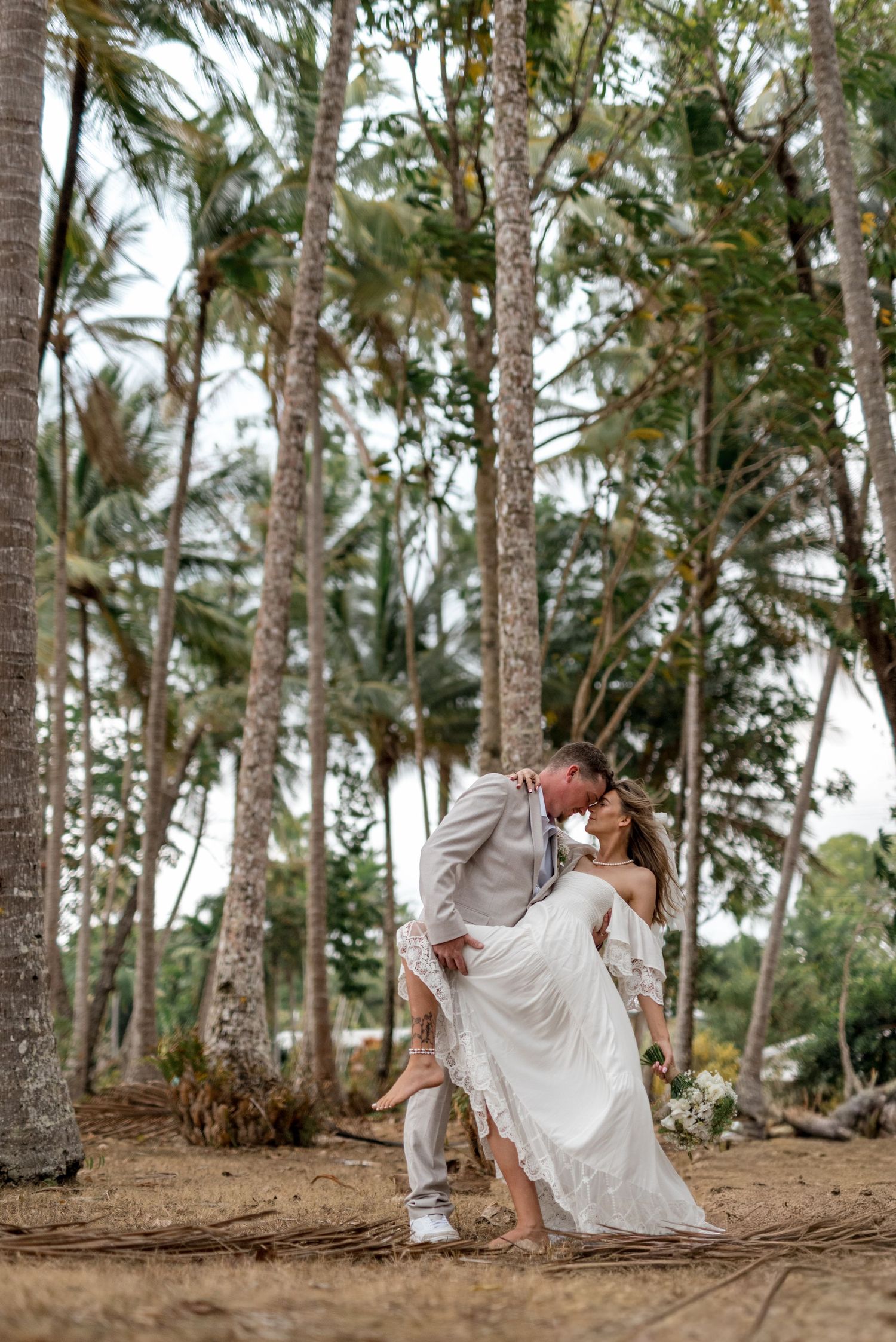 Bride and groom sharing a romantic dip kiss among tall palm trees on a tropical beach wedding day.