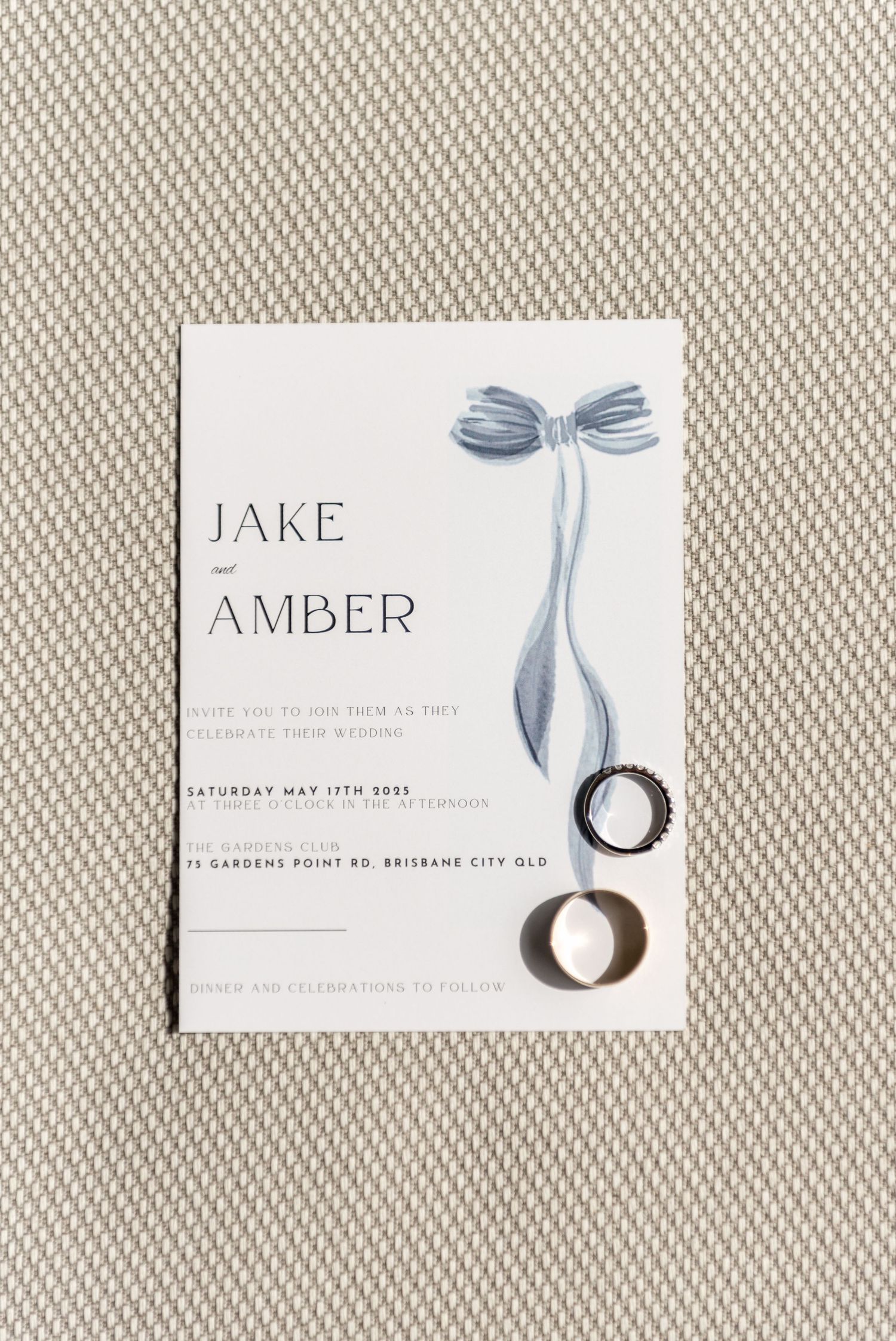 Wedding invitation for Jake and Amber with blue bow illustration and gold rings.