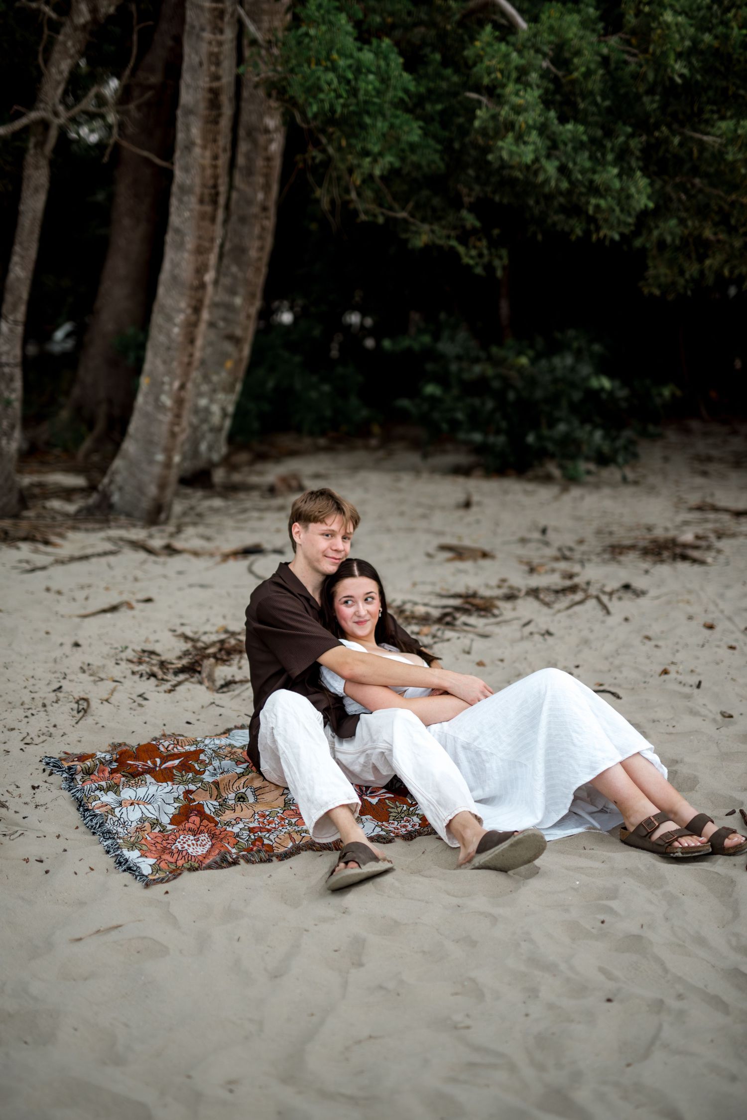 Couple sits closely on bohemian blanket on sandy beach surrounded by trees.