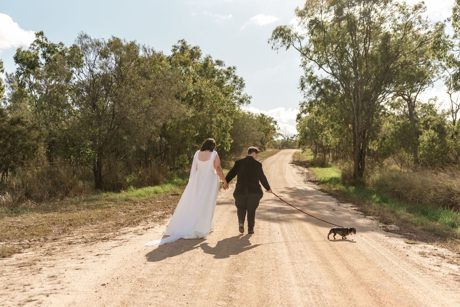 Bride and groom walk their small black dog down a tree-lined sandy country dirt road.
