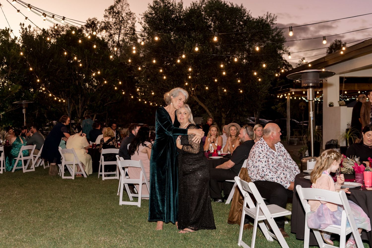 Two women in elegant gowns walk through outdoor wedding reception seating area at dusk.