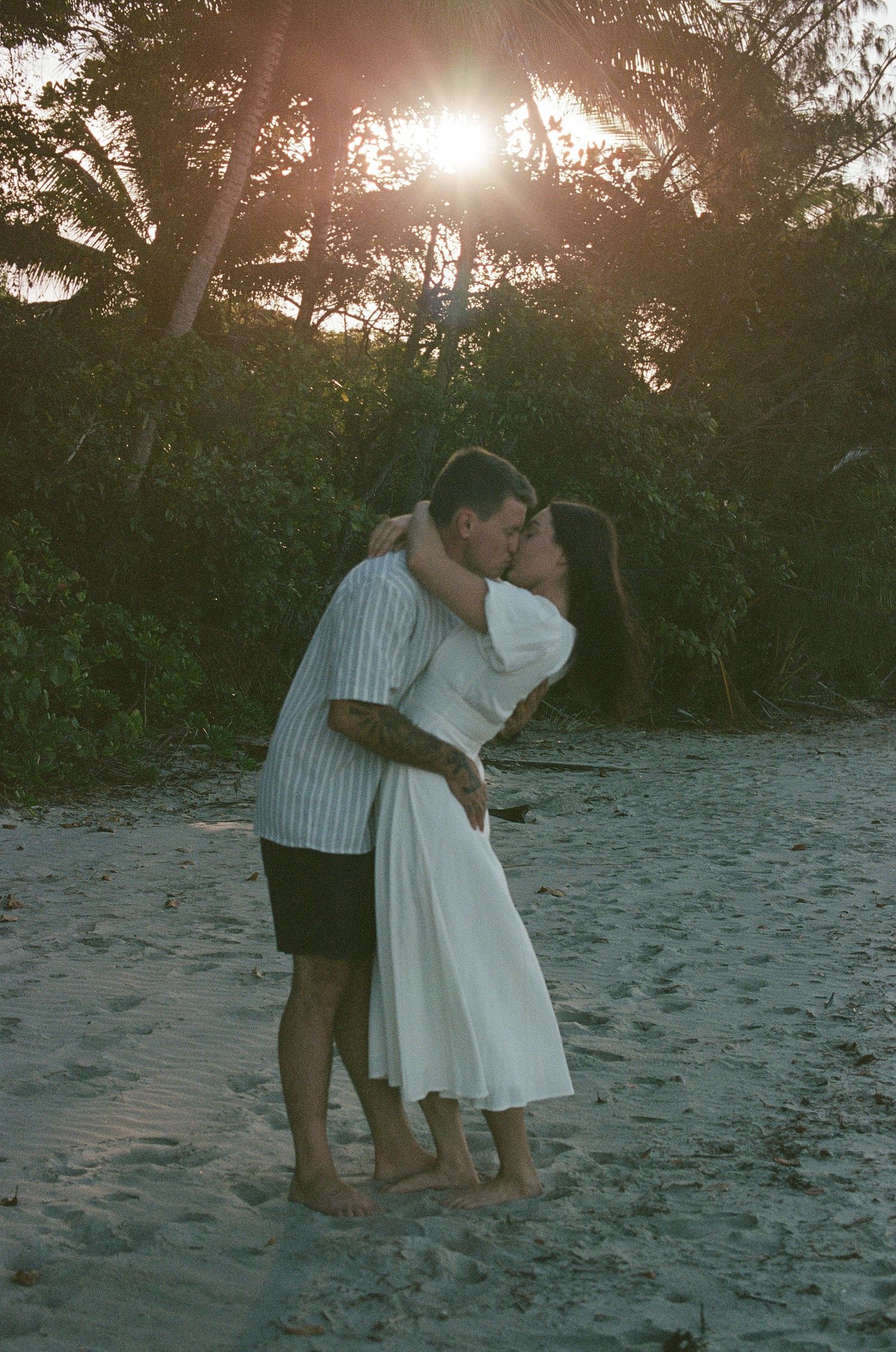 Couple embracing and kissing on a sandy beach with sunlight streaming through lush tropical trees.