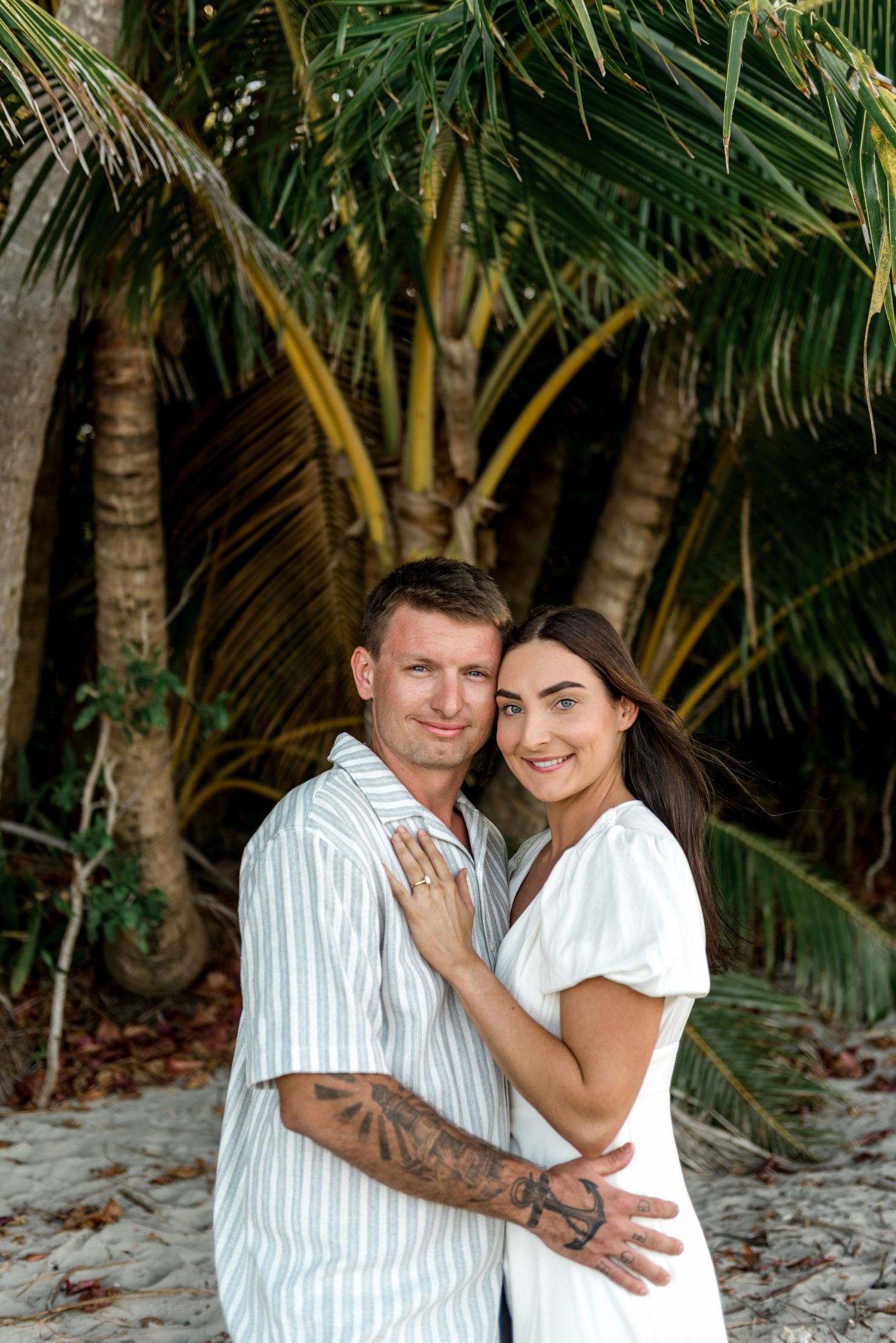 Smiling engaged couple embracing on white sandy beach with lush tropical palm trees behind them.