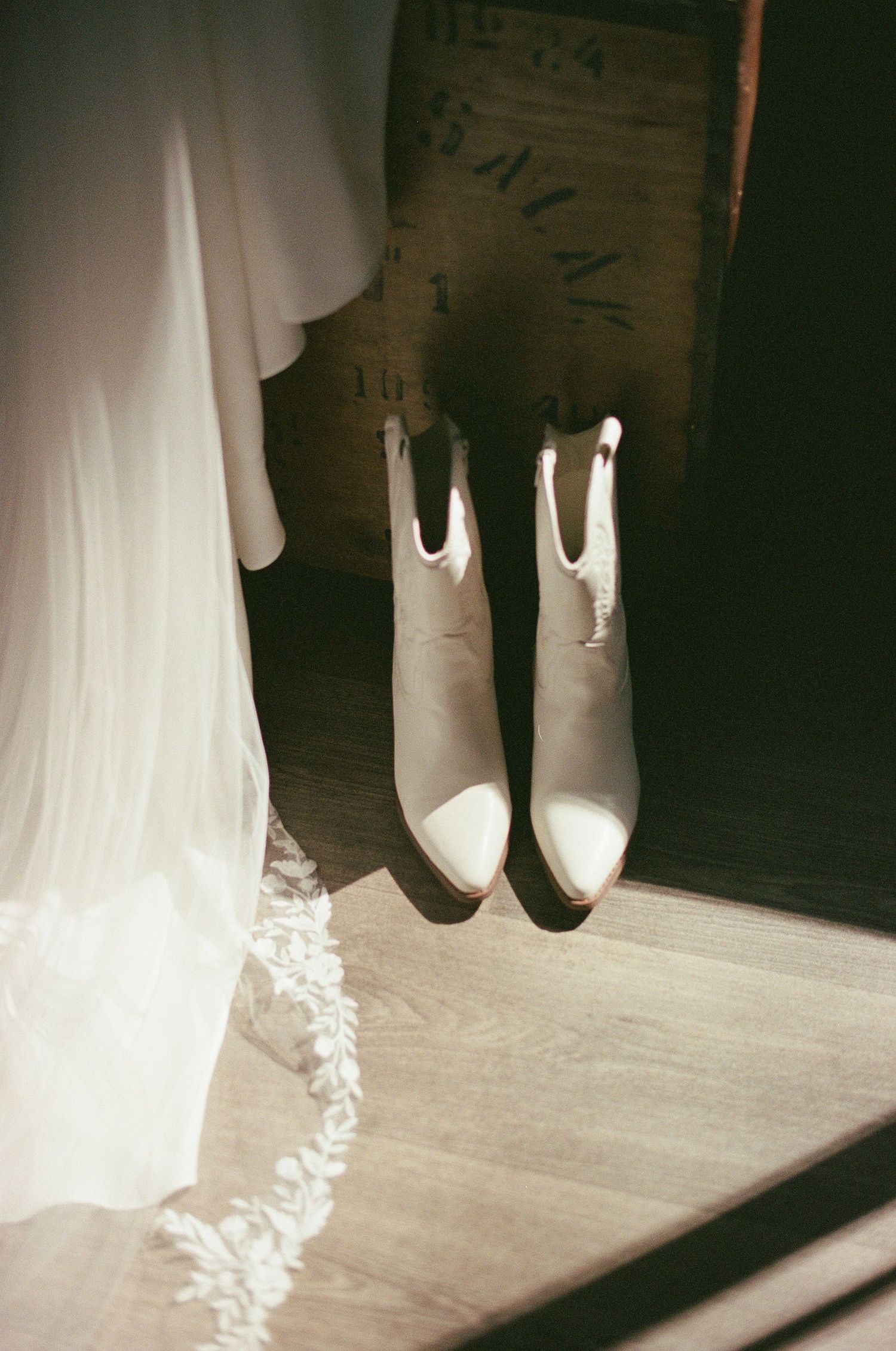 White bridal cowboy boots and lace veil displayed in sunlit rustic wooden setting.