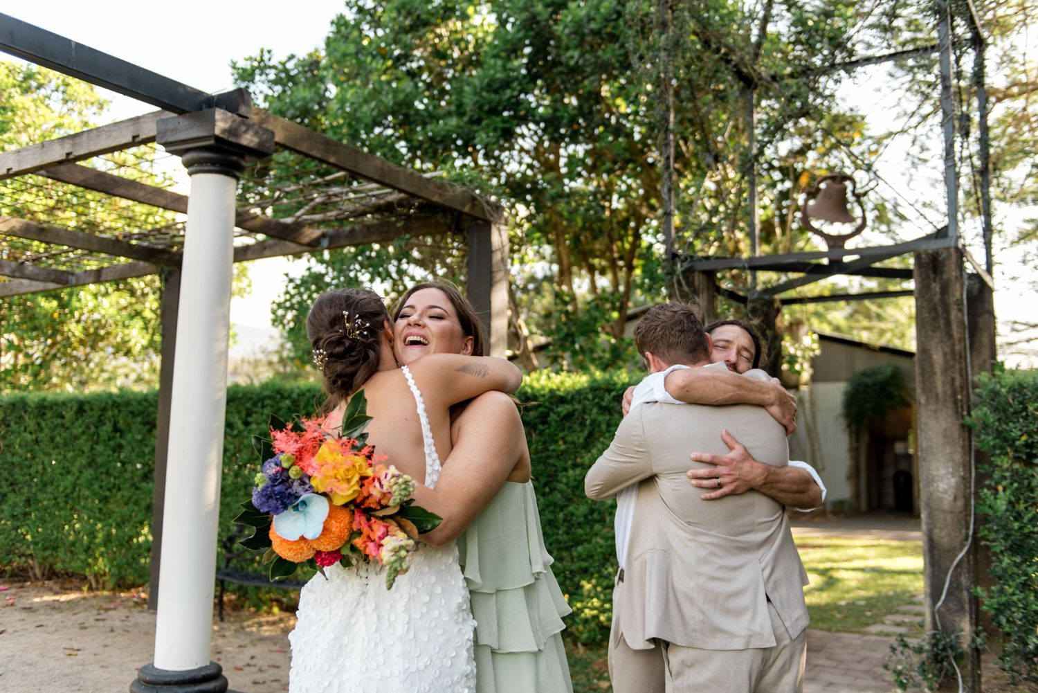 Bride and bridesmaid embrace guests with hugs under rustic pergola at outdoor wedding.