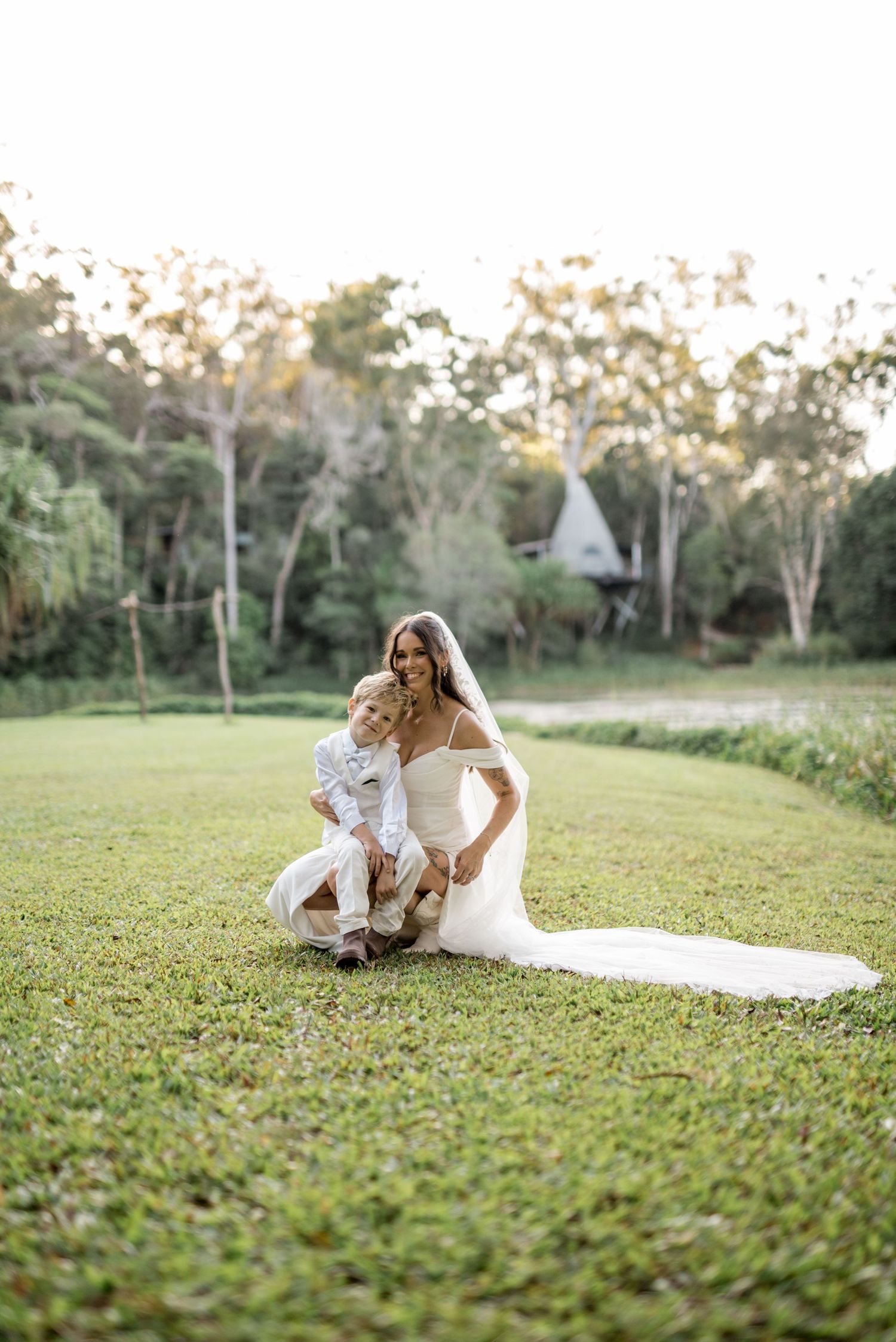 Bride in white gown and veil crouches on green lawn hugging a young child at sunset.