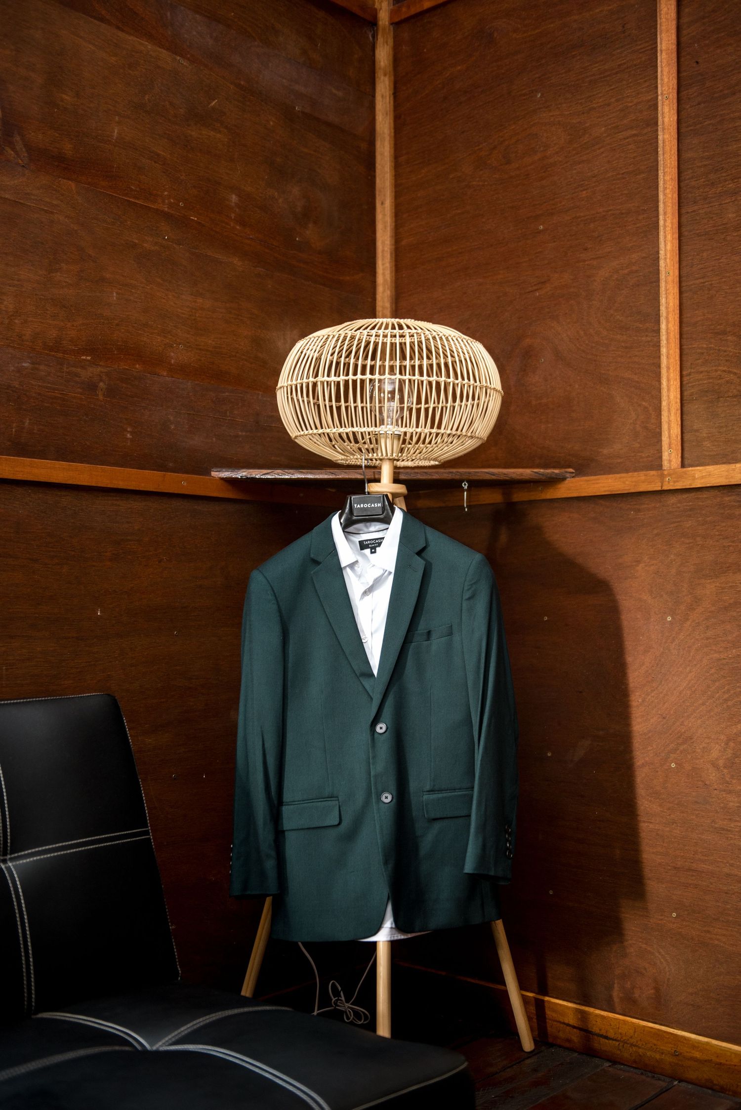 Dark green suit jacket hanging on a rattan lamp stand in a rustic wooden paneled room.