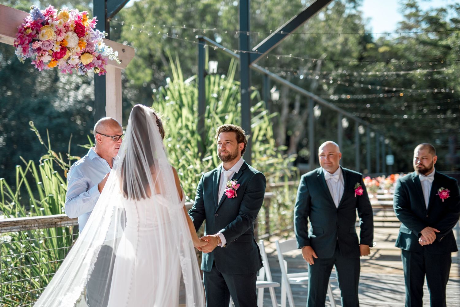 Groom smiling at bride during outdoor wedding ceremony on a wooden deck with groomsmen standing alongside in dark suits.