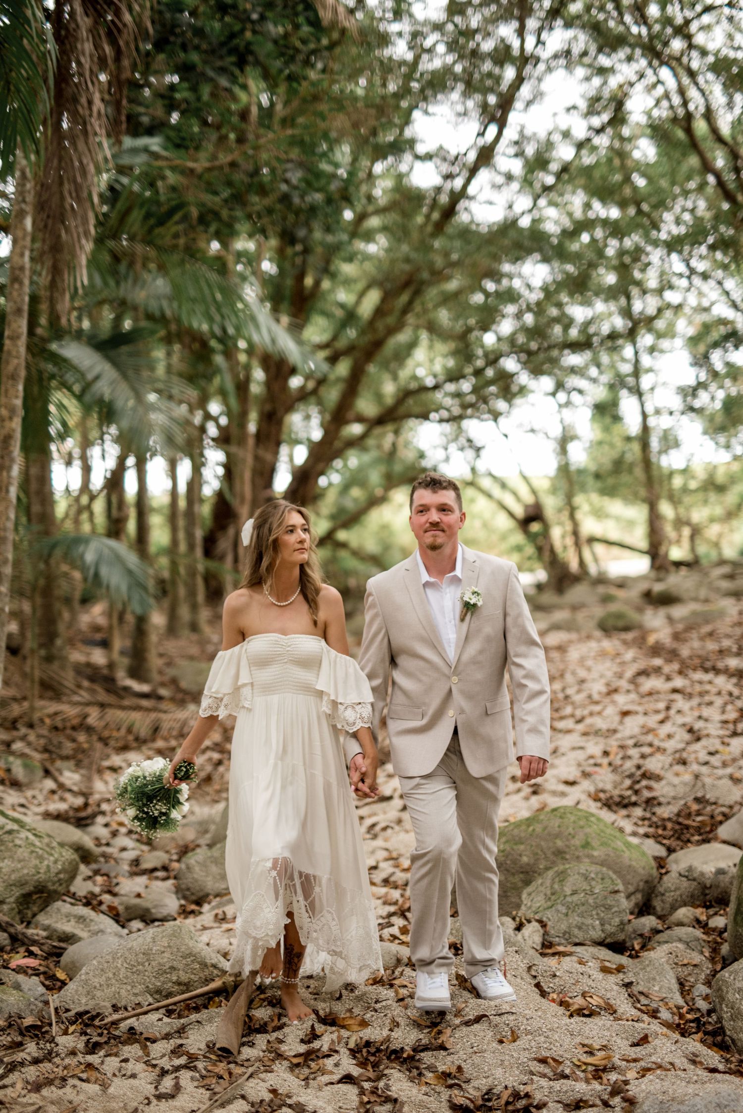 Bride and groom walking hand-in-hand through rocky forest, bride in boho white dress holding bouquet, groom in beige suit.