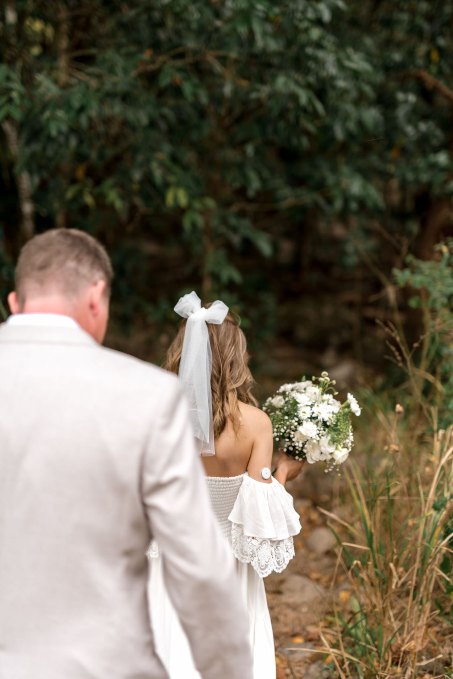 Bride holding white bouquet with bow veil walking beside groom in lush forest.