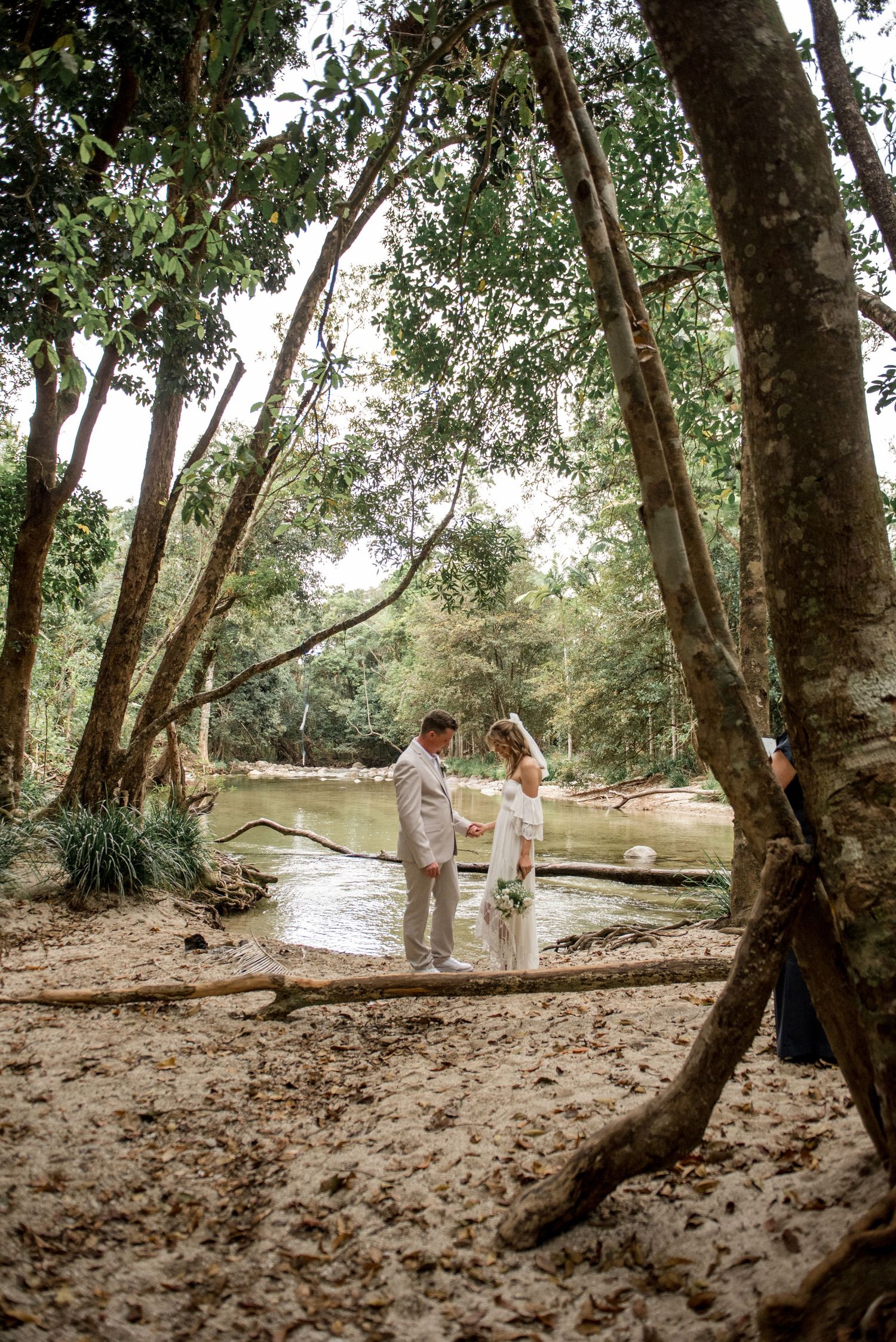 Wedding couple holds hands between trees beside a tranquil jungle river.