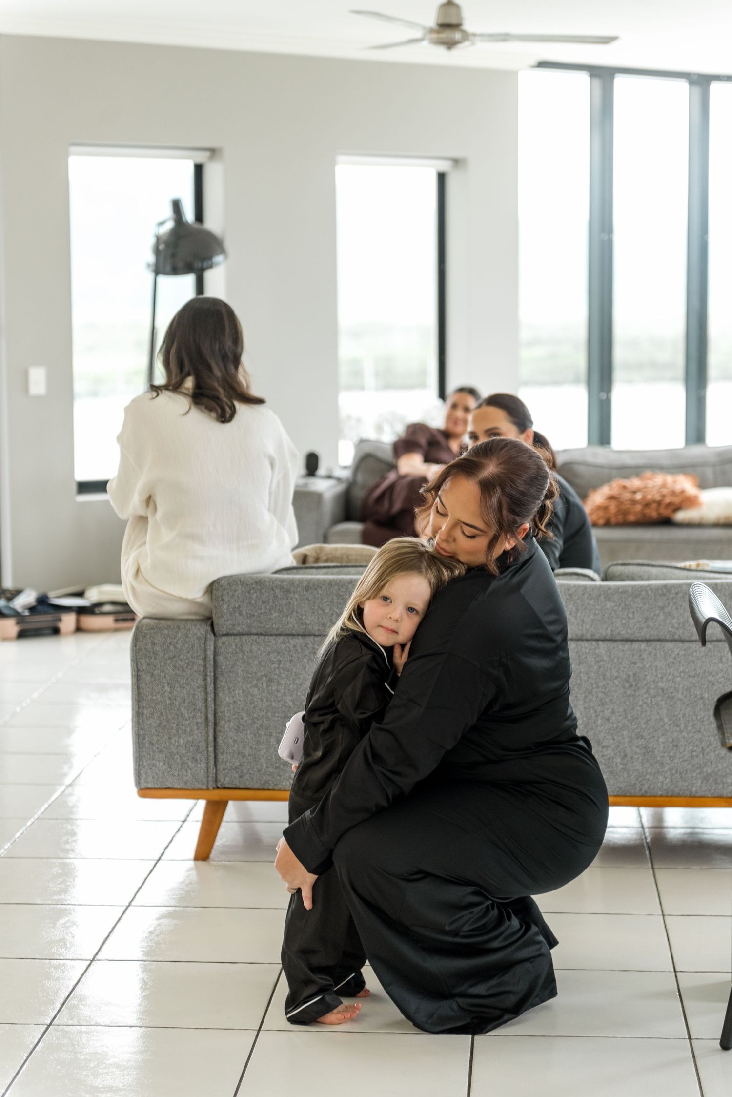 Woman in black outfit crouching to hug a young child in a modern living room during wedding preparations.