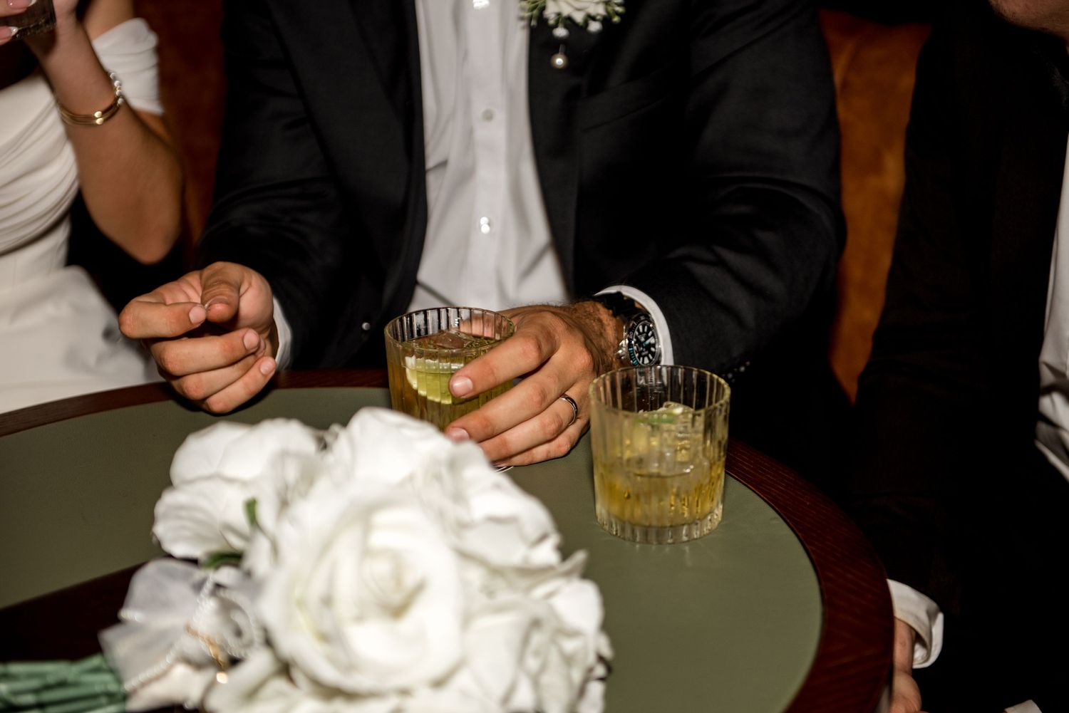 Groom holds whiskey glasses near a white rose bridal bouquet on a round table.