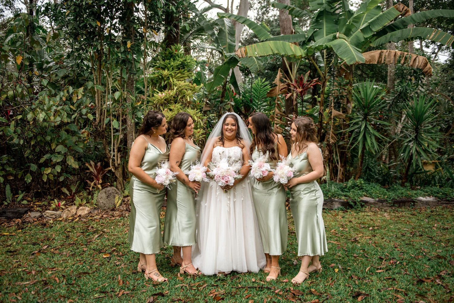 Bride with veil stands with four bridesmaids in sage satin dresses holding pink bouquets in garden.