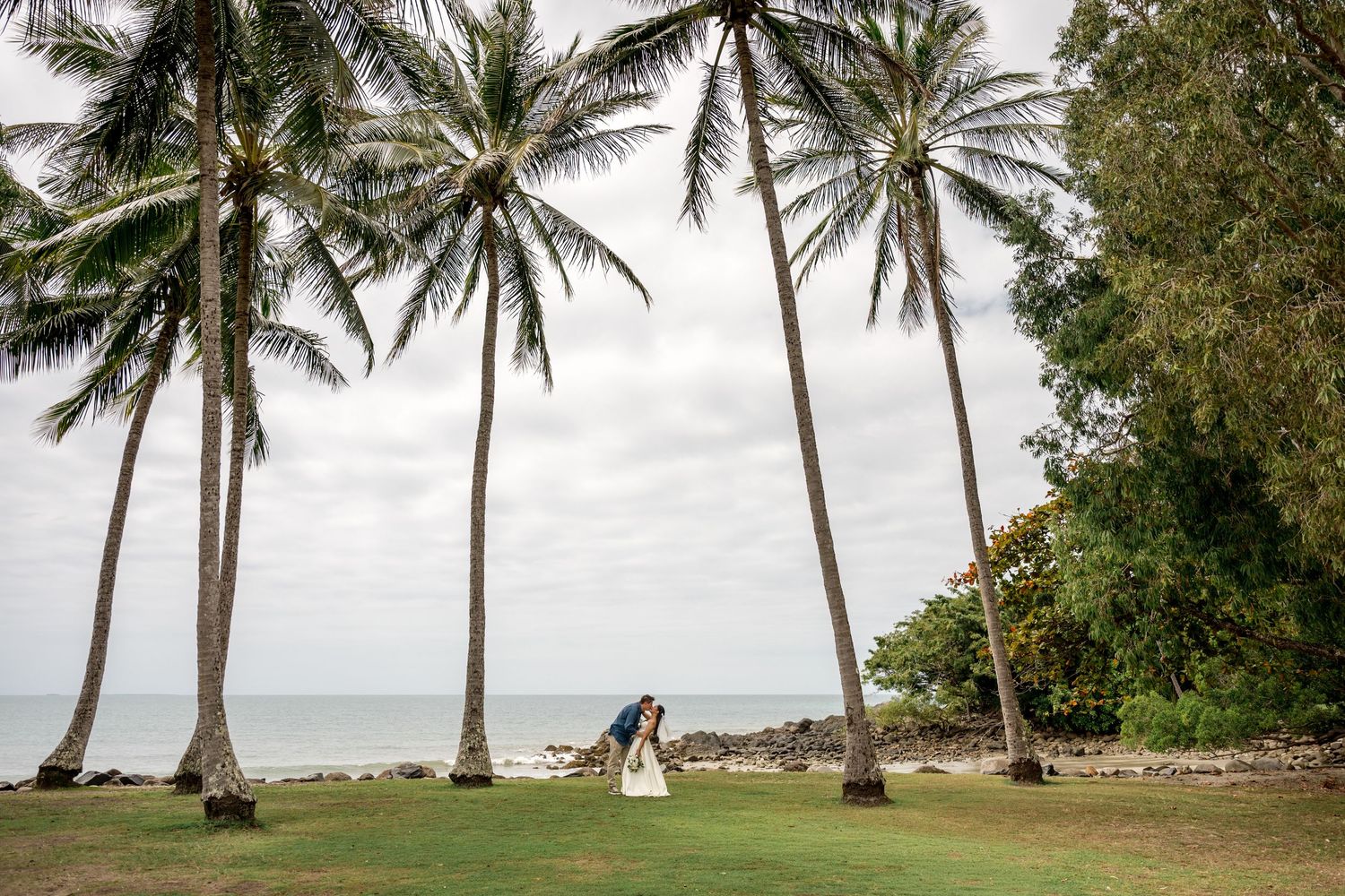 Newlywed couple shares tender moment on tropical lawn surrounded by tall swaying palm trees at Little Cove, Port Douglas.