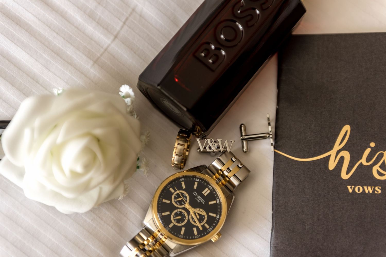 Groom's wedding details flat lay with gold watch, white rose, cologne, cufflinks, and vow book on white linen.