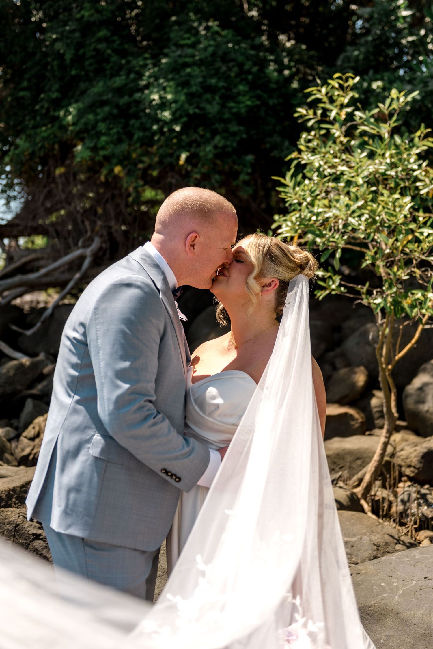 Bride and groom sharing a romantic kiss near rocky shoreline with lush green trees backdrop.