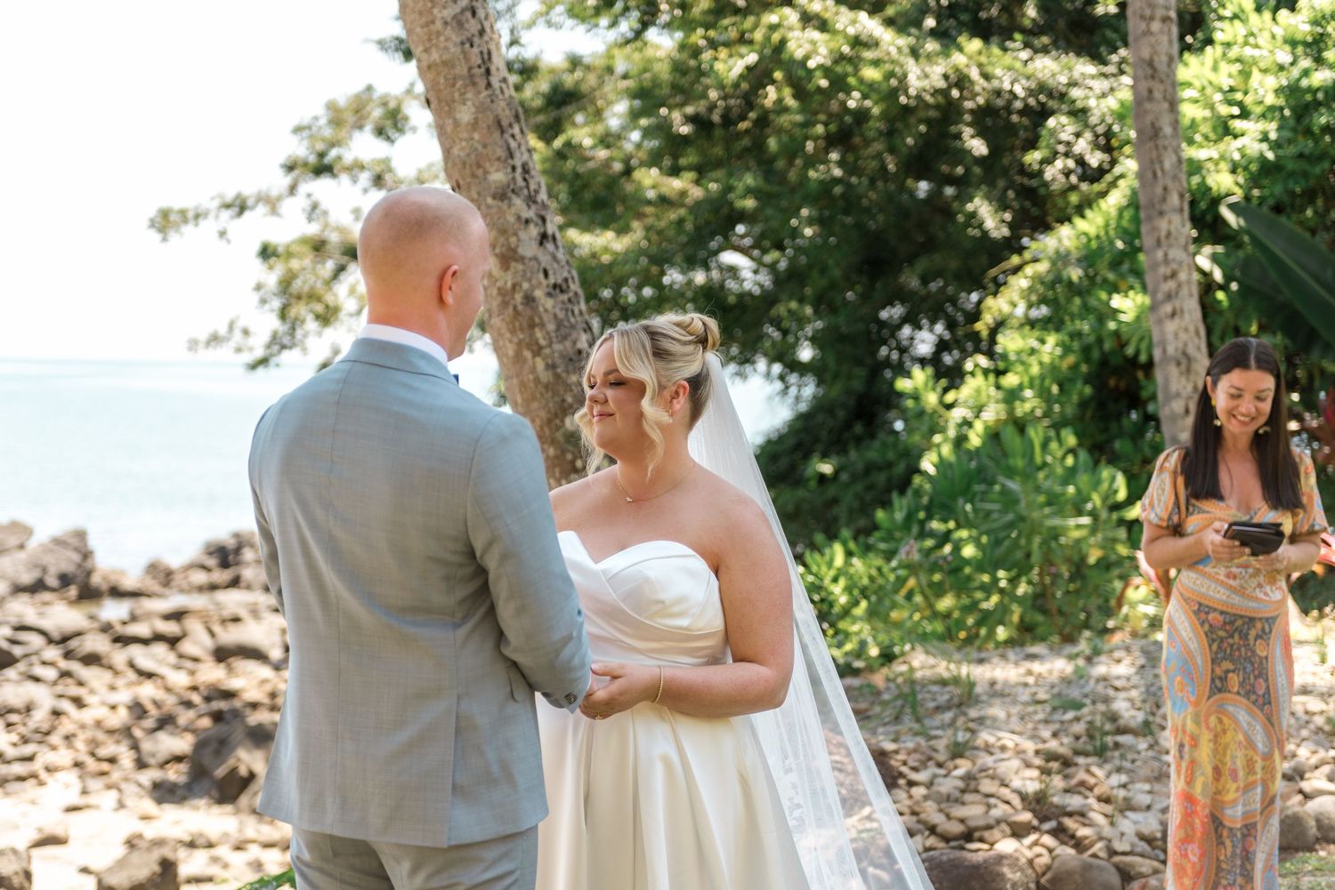 Bride in strapless gown holds grooms hands during intimate beachside wedding ceremony with officiant.