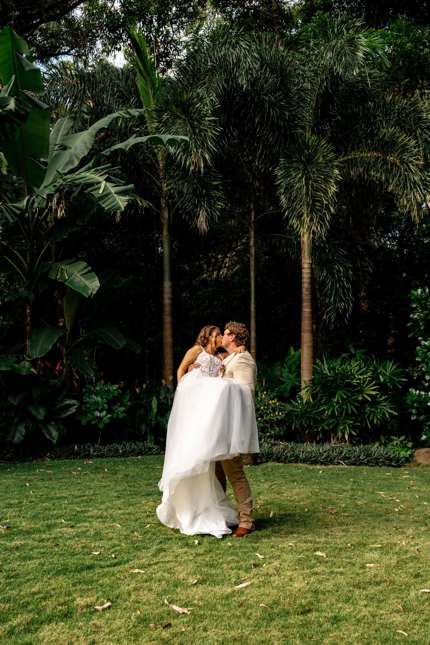 Newlyweds sharing passionate kiss on green lawn surrounded by tropical palms and lush garden foliage.
