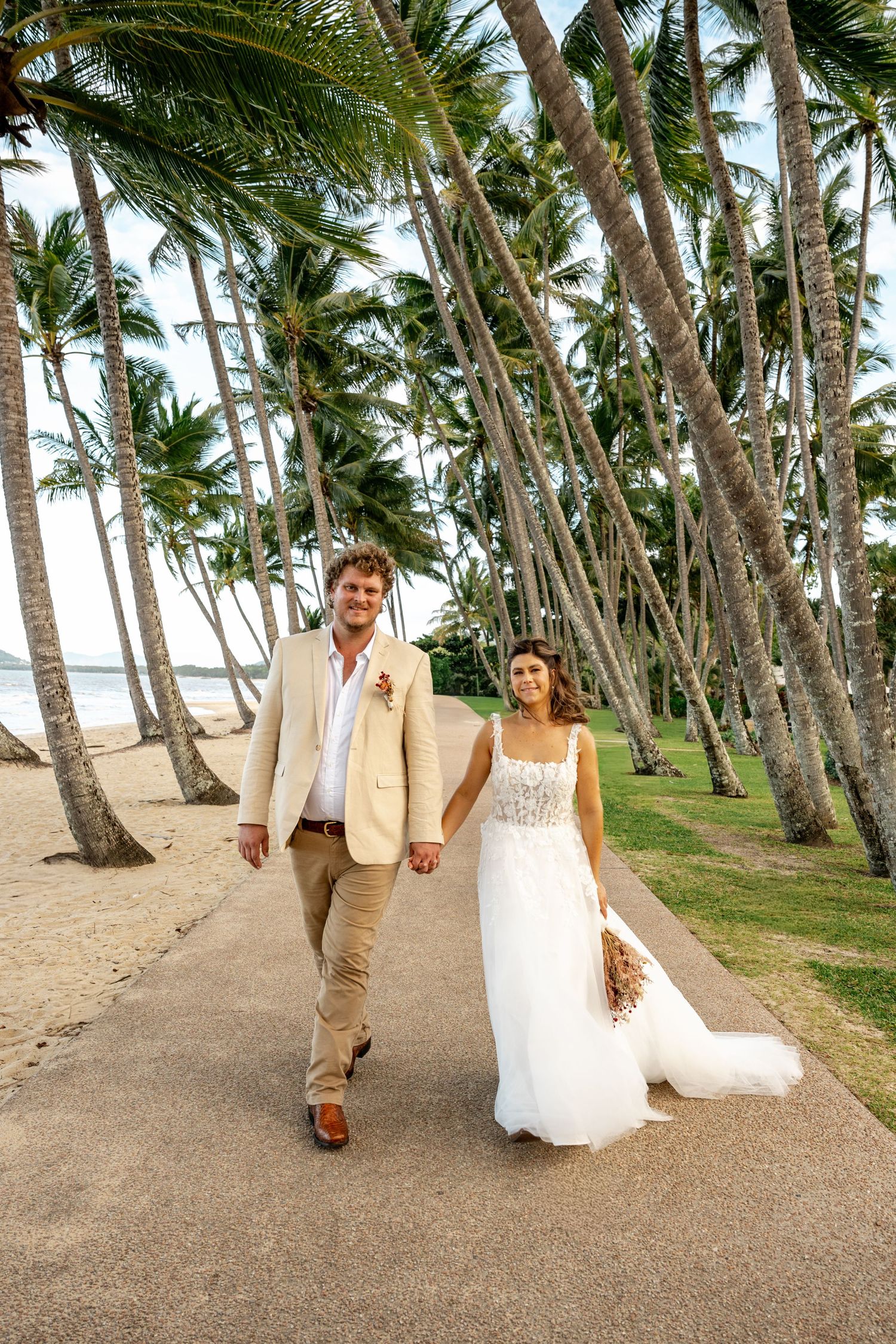 Smiling newlyweds in wedding attire stroll hand-in-hand along a tropical palm-lined beach walkway.