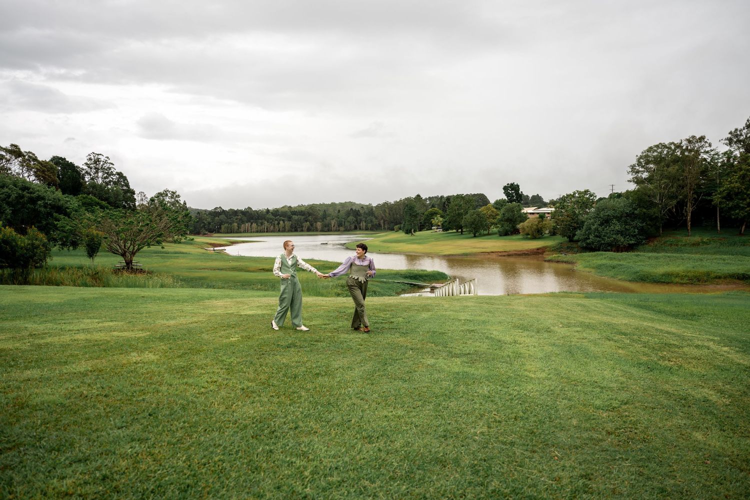 Couple in green outfits holding hands on a wide green lawn with a scenic pond backdrop.