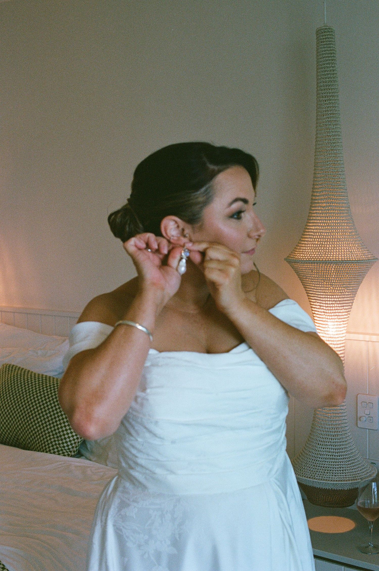 Bride in white off-shoulder wedding dress putting on drop earrings beside a woven lamp in warm light.