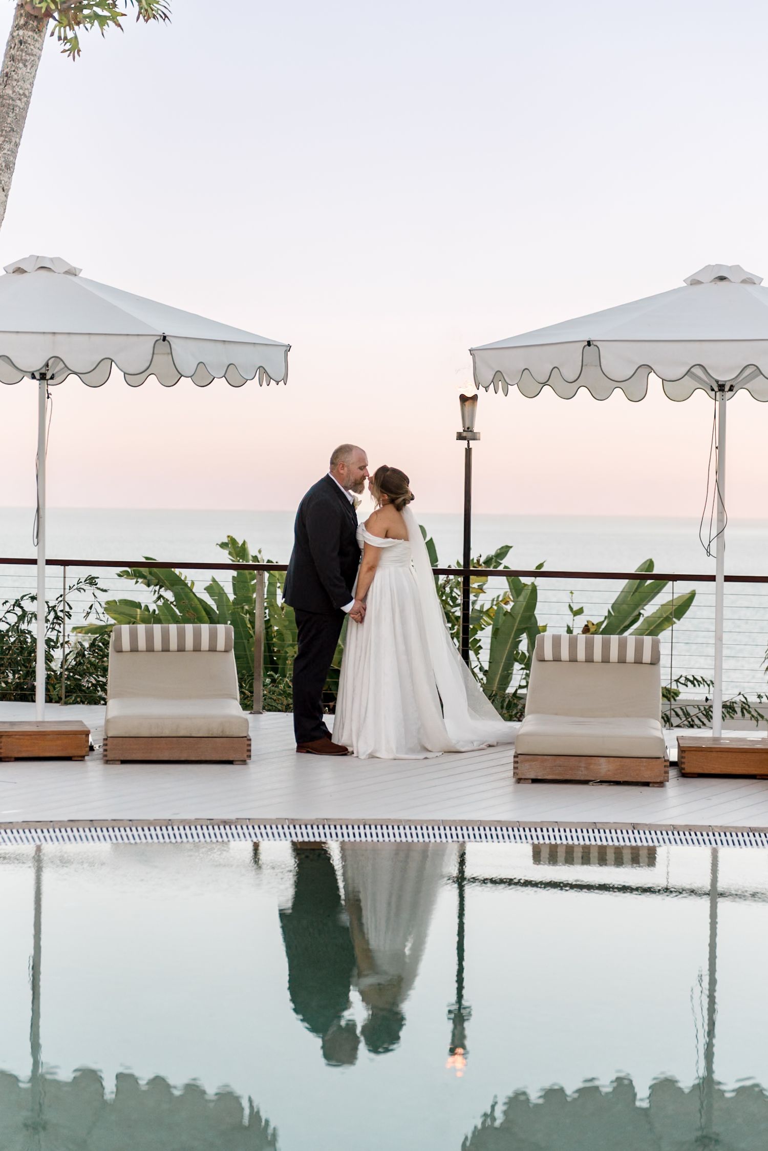 Bride and groom embrace poolside at twilight with their reflection visible in the calm resort pool water.