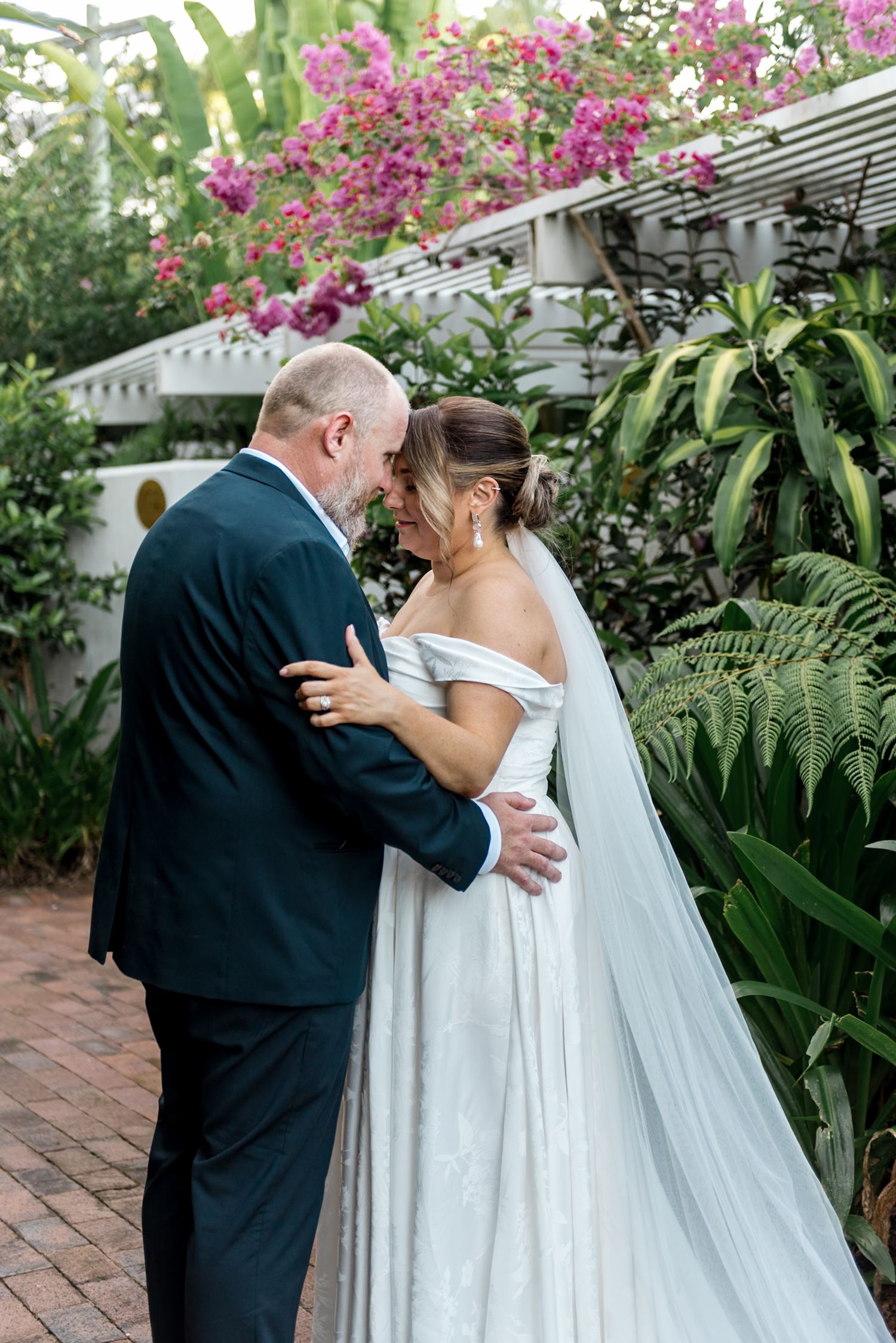 Bride in off-shoulder gown and groom embrace in garden with pink bougainvillea blooms.