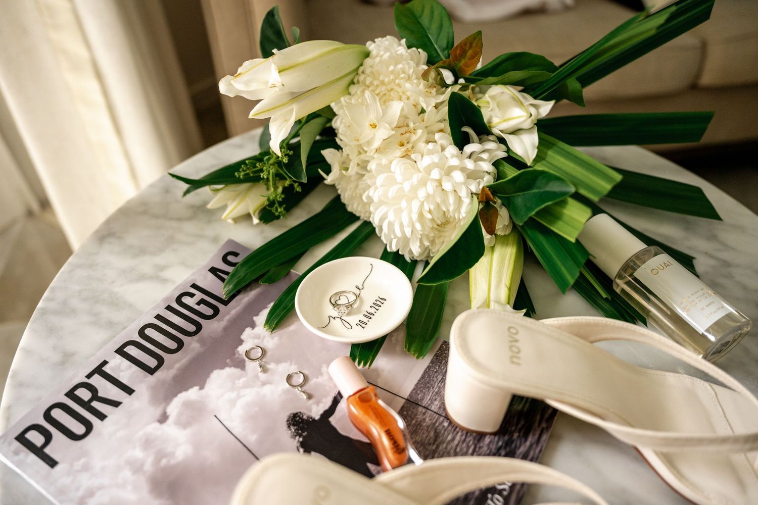 Overhead view of white bridal sandals, ring dish, and white floral bouquet on a Port Douglas magazine.