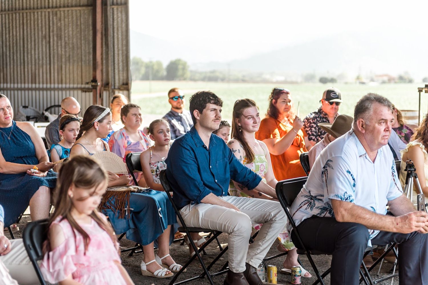 Multigenerational wedding guests seated in rustic barn ceremony venue with scenic green fields visible in background.