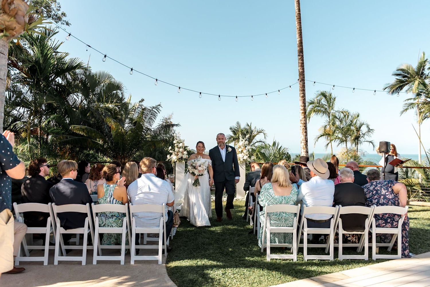 Outdoor tropical wedding ceremony with guests seated in white chairs under string lights with palm trees and ocean views.