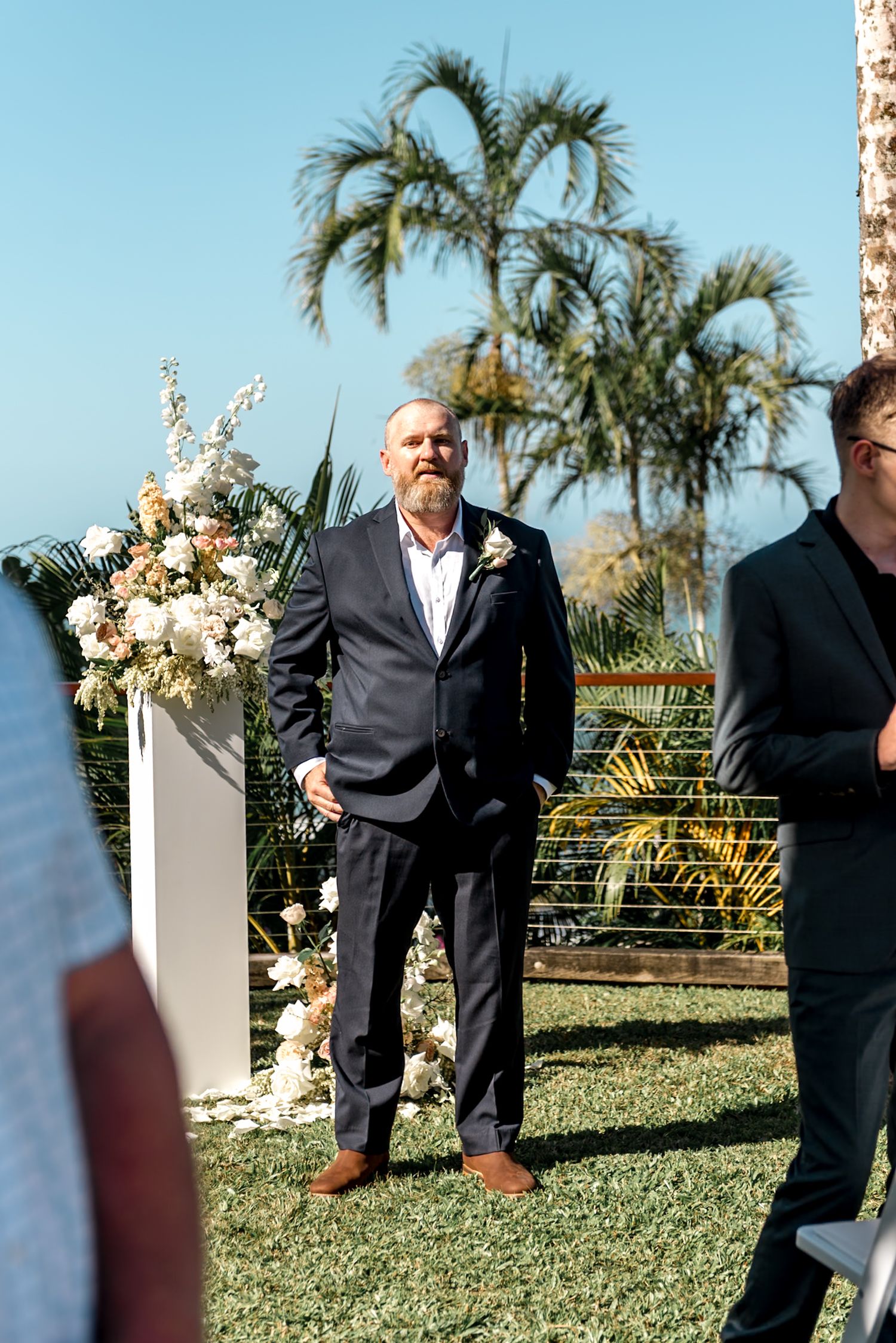 Bearded groom in navy suit stands at outdoor tropical wedding ceremony beside white floral arrangements.