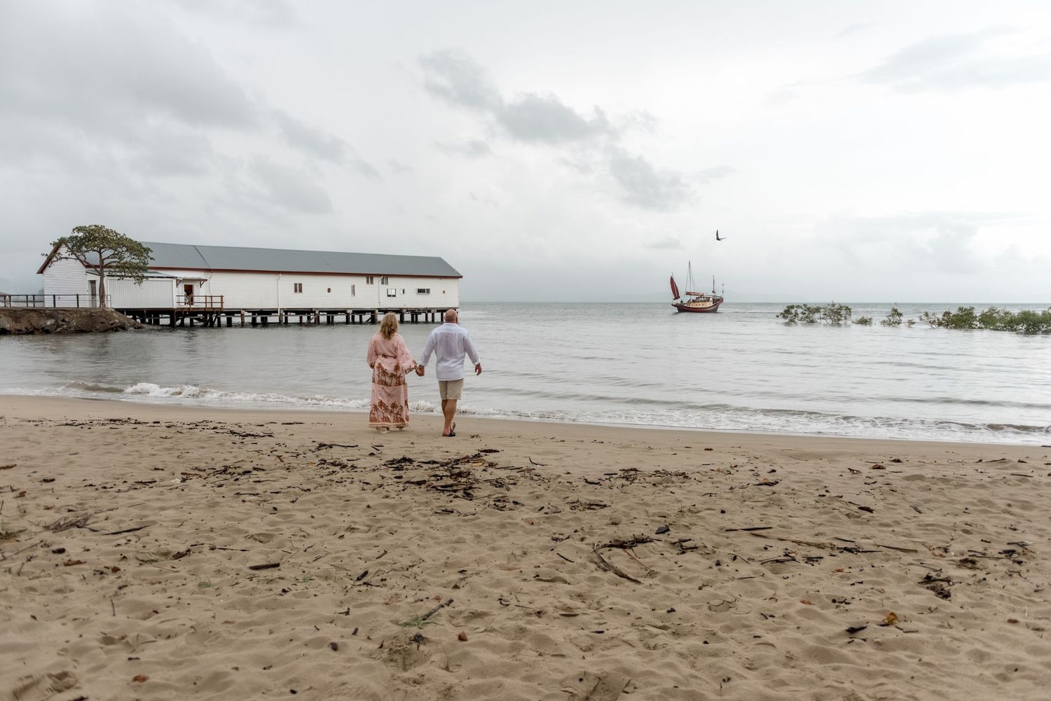 Couple walking hand in hand on a sandy beach toward a historic pier on a cloudy day.